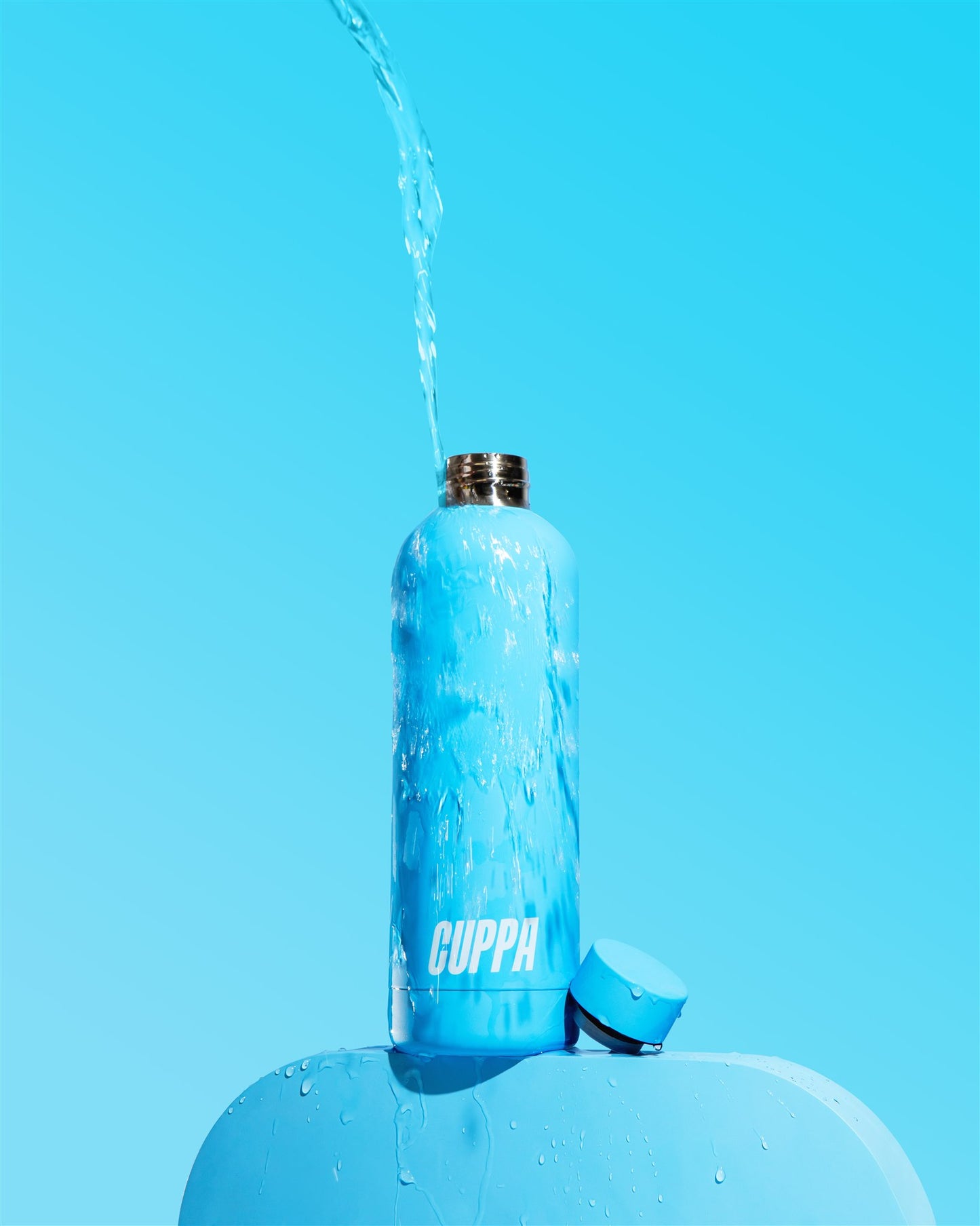 Bright Blue Cuppa Water Bottle with water pouring onto it from above on a matching blue background